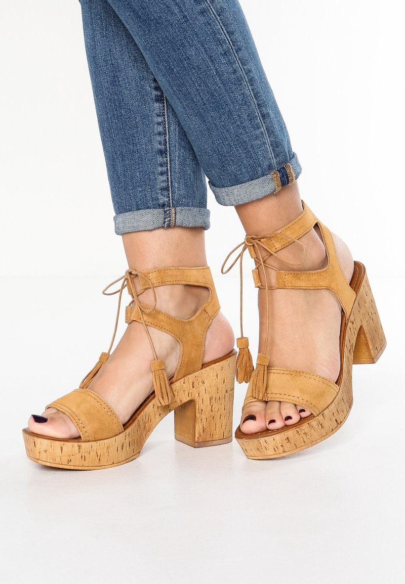 Pieds en jean bleu retroussé portant des sandales compensées en suède beige avec talons blocs et lacets à pampilles à la cheville sur fond blanc.