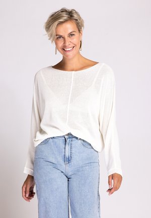 Smiling woman with short blonde hair wearing a white loose sweater and light blue high-waisted jeans against a plain background.