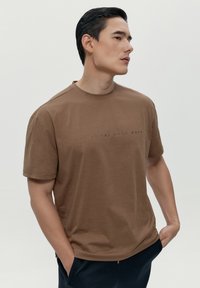 Hnědé tričko s krátkým rukávem a kulatým výstřihem, na přední straně s jemným grafickým textem. Tkanina se zdá být měkká s hladkým povrchem.