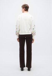 Λευκό φούτερ με μανίκια σε λουλουδάτο σχέδιο, συνδυασμένο με καφέ παντελόνι καμπάνα. Πίσω όψη χωρίς σχέδιο, με απαλό ύφασμα και εφαρμοστή σιλουέτα.