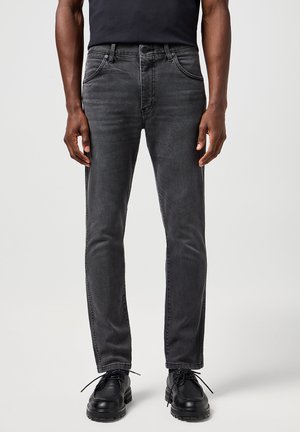 Άνδρας που φοράει στενά, ξεβαμμένα μαύρα τζιν και μαύρα δερμάτινα παπούτσια με κορδόνια, στέκεται μπροστά σε απλό φωτεινό φόντο.