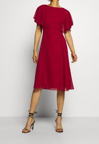 Robe rouge mi-longue avec manches courtes transparentes volantées, taille cintrée, jupe fluide, associée à des sandales à talons hauts noires à brides.