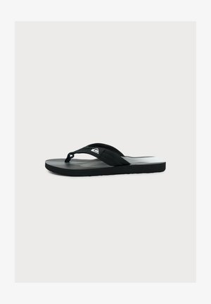 Čierno-šedá šľapka typu flip-flop so širokými textilnými remienkami a malým bielym logom na remienku, zobrazená zboku na bielom pozadí.