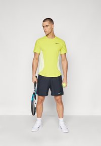 Mandlig atletisk påklædning med en lys gul kortærmet skjorte, sorte shorts og hvide sneakers. Holder en tennisketsjer og en bold.
