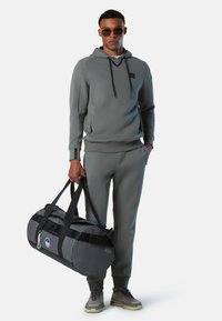 Grauer Kapuzenpullover mit Kordelzügen, passende Jogginghose und eine Sporttasche. Die Tasche hat ein rundes Design und ein 45L-Etikett. Graue und schwarze Sneaker.