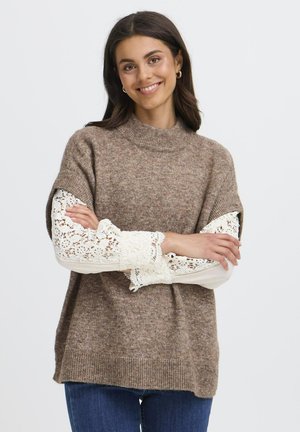 Junge Frau mit dunklen Haaren, die einen braunen Strickpullover über einem weißen Spitzenoberteil mit langen Ärmeln trägt, lächelnd mit verschränkten Armen.