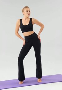 Un crop top noir et un pantalon de yoga évasé en tissu extensible, sur fond gris clair. Le mannequin pose sur un tapis de yoga violet.