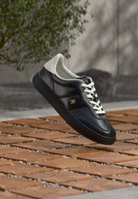 Μαύρο δερμάτινο αθλητικό παπούτσι με λεία επιφάνεια, λευκές λεπτομέρειες και επίπεδα κορδόνια, με ένα μικρό χρυσό έμβλημα στο πλάι.