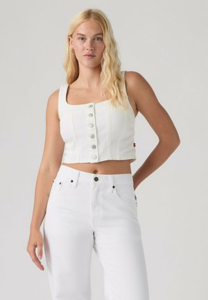 Blondynka w białym, bezrękawnym crop topie zapinanym na guziki i białych jeansach z wysokim stanem, stojąca na jednolitym tle.