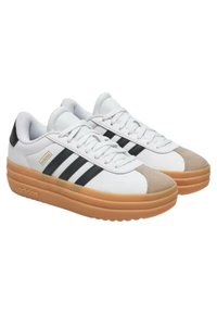 Zapatillas blancas con tres rayas negras, puntera de ante color marrón claro, lengüeta acolchada y suela de goma texturizada. Logo de la marca en el lateral.