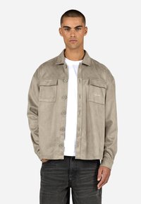 Beige velúr dzseki gombos elülső résszel, két mellzsebbel és diszkrét logó hímzéssel. Fehér ing és sötét farmer fölött viselt.