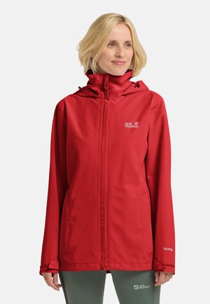 Frau trägt eine rote Kapuzen-Regenjacke mit Jack Wolfskin-Logo und grüne Hose vor einem einfarbigen weißen Hintergrund.