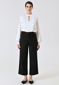 Blusa bianca con scollatura a keyhole e maniche lunghe, abbinata a pantaloni neri a vita alta e a gamba larga, insieme a ballerine nere con fiocco.