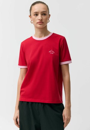 Kobieta ubrana w czerwoną koszulkę z różowym kołnierzykiem i mankietami oraz białym napisem „True Love Club” na piersi, w połączeniu z czarnymi spodniami w prążki.