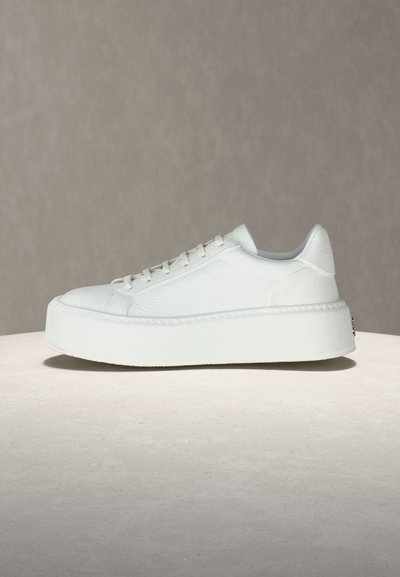 Baskets à plateforme en cuir blanc avec semelle épaisse et lacets, présentées sur une surface neutre sur fond beige.