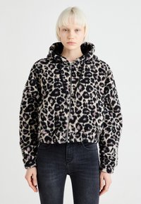 Jakna s kapuljačom u leopard uzorku izrađena od mekane, strukturirane tkanine. Ima zatvaranje na zip, crne detalje i skraćeni dizajn. Uparena s tamnim trapericama.