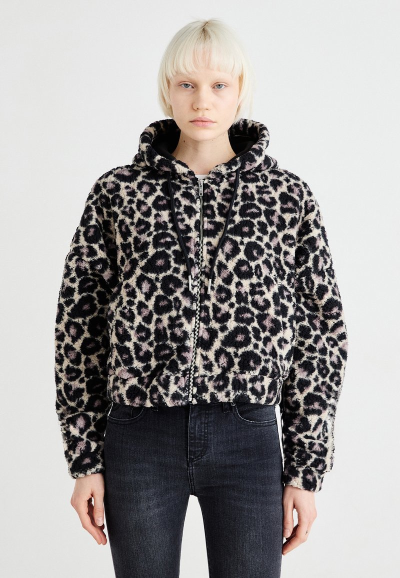 Jakna s kapuljačom u leopard uzorku izrađena od mekane, strukturirane tkanine. Ima zatvaranje na zip, crne detalje i skraćeni dizajn. Uparena s tamnim trapericama.