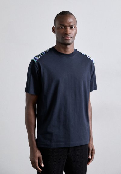 T-shirt à manches courtes bleu marine en coton, avec des accents de motifs en zigzag verts et violets sur les épaules.
