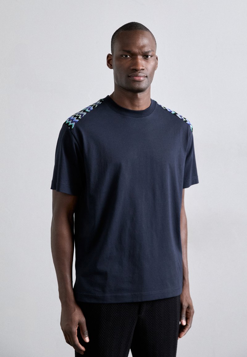 T-shirt à manches courtes bleu marine en coton, avec des accents de motifs en zigzag verts et violets sur les épaules.