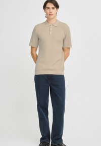 Beige bordázott pólóing rövid ujjakkal, három gomb a gallérnál, sötétkék farmernadrággal párosítva. Egyszerű és texturált dizájn.