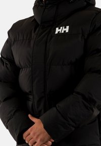 Giubbotto imbottito nero con colletto alto, chiusura con zip e logo ricamato bianco. Il tessuto sembra liscio e caldo, con un design strutturato.