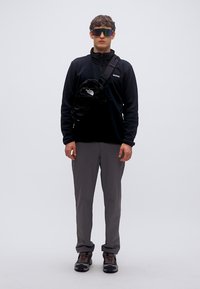 Μαύρο φλις μπουφάν με φερμουάρ, γκρίζο παντελόνι και μαύρη τσάντα χιαστί; το μοντέλο φοράει ανακλαστικά γυαλιά ηλίου και ορειβατικά παπούτσια. Μινιμαλιστικός σχεδιασμός.