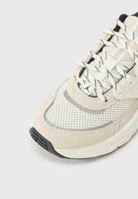 Μπεζ αθλητικό παπούτσι με επάνω μέρος από δίχτυ, σουέντ λεπτομέρειες και μαύρη λαστιχένια σόλα. Τα κορδόνια έχουν ασπρόμαυρο μοτίβο, με λογότυπο στη πλευρά.