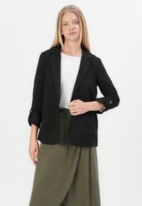 Zwarte blazer met twee voordekselzakken, opgerolde manchetten en een gestructureerd ontwerp. Gecombineerd met een olijfgroene rok, wat een gelaagd uiterlijk creëert.