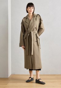 Trench coat lungo color verde oliva con design doppiopetto, vita con cintura, grandi tasche e ampi revers. Indossato con calzature nere.