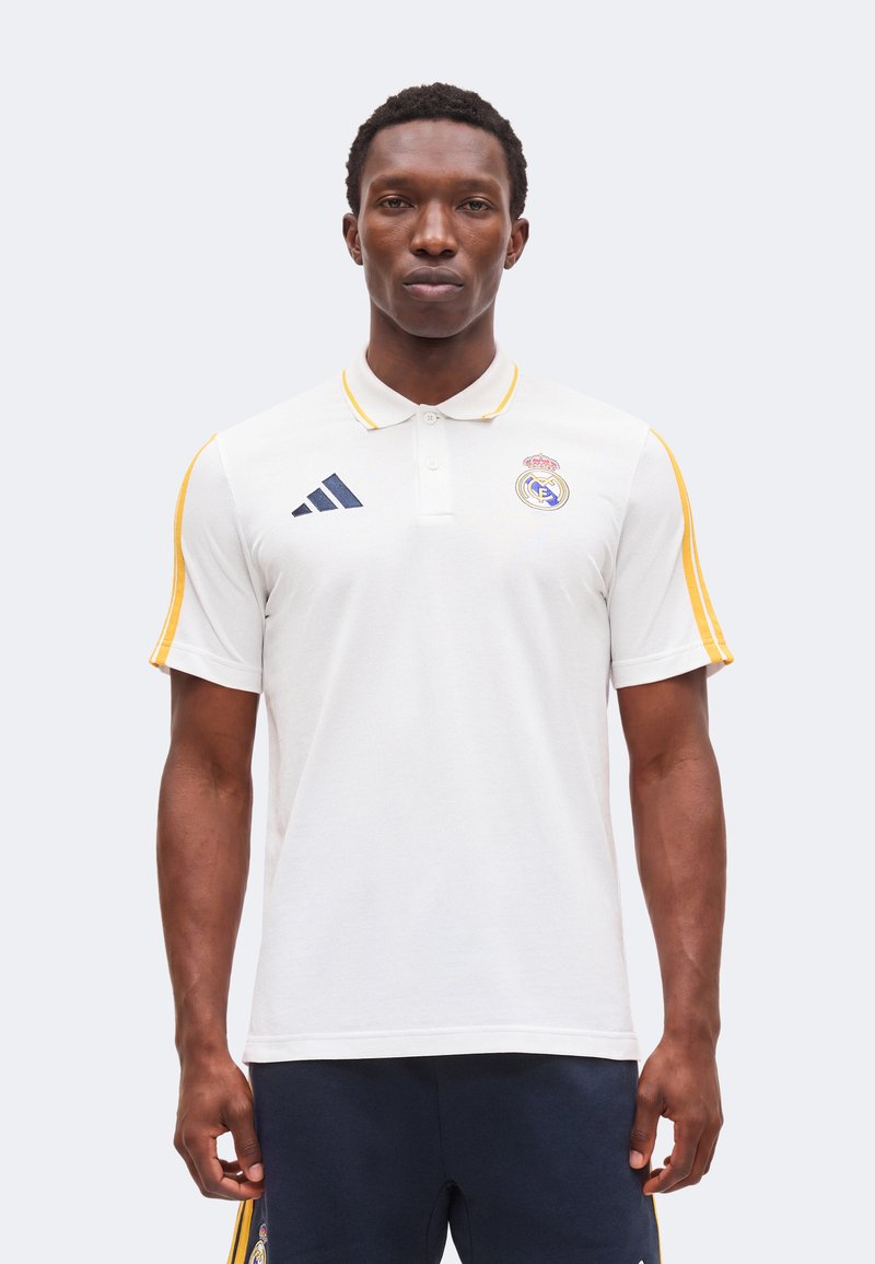 Homme debout face à l'avant portant un polo blanc avec des rayures jaunes et les logos du Real Madrid et Adidas.