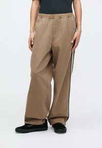 Oseba, ki nosi široke bež hlače s črno-belimi stranskimi črtami in črne superge, stoji pred enostavnim ozadjem.