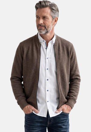 Mann mit grauen Haaren und Bart trägt eine braune Zip-up-Jacke über einem weißen Hemd mit Knöpfen, steht mit den Händen in den Taschen und schaut nach links.