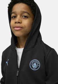 Crni dukserica na zip sivi Puma logo, s plavim grbom Manchester Cityja na lijevoj strani prsnog dijela. Mekana, glatka tkanina s kapuljačom.