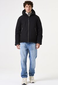 Chaqueta negra aislante con capucha, cierre de cremallera y bolsillos laterales, combinada con jeans de mezclilla azul claro y zapatillas en tonos neutros. Textura suave.