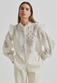 White button-up blouse made of textured fabric with cut-out circles and vertical lines, featuring ruffled shoulders and loose sleeves.