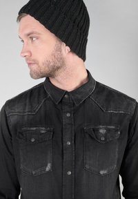 Camicia in denim nera con due tasche sul petto, chiusura a bottoni e dettagli sbiaditi. Berretto nero a coste; modello rivolto leggermente a sinistra.