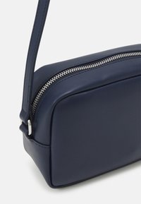 Borsa a tracolla in pelle blu navy con forma rettangolare, caratterizzata da una cerniera in argento e una cintura slanciata regolabile. Texture liscia con dettagli minimi.