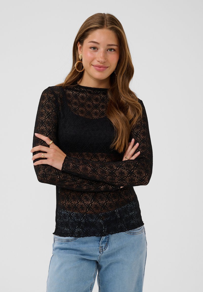 Jeune femme aux longs cheveux bruns portant un haut noir en dentelle à manches longues et un jean bleu clair, bras croisés, souriant doucement.