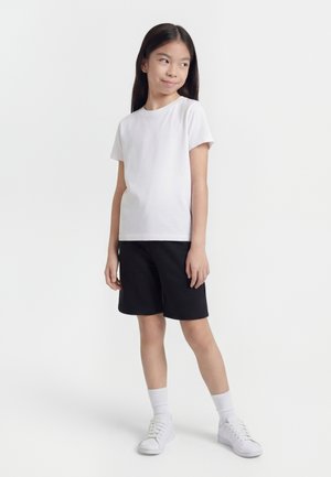 Ung jente som står sidelengs iført en ensfarget hvit t-skjorte, svarte shorts, hvite sokker og hvite joggesko på hvit bakgrunn.