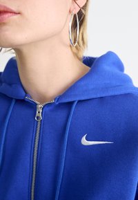 Zils kapuci džemperis ar rāvējslēdzēju, izgatavots no mīksta auduma, ar baltu Nike logo kreisajā krūtīs un ar auklu uz kapuces. Redzami sudraba riņķa auskari.