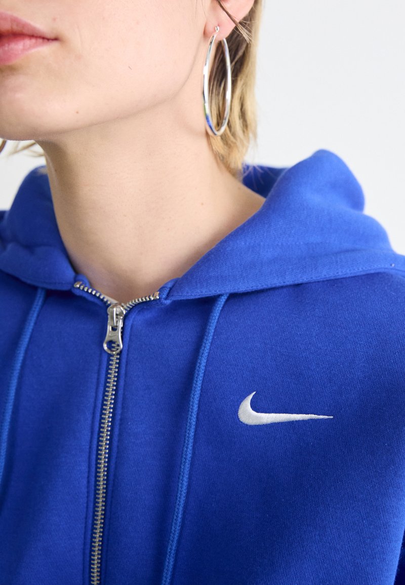 Sudadera azul de cremallera hecha de tela suave con un logo blanco de Nike en el lado izquierdo del pecho y una capucha con cordón. Se pueden ver pendientes de aro plateados.