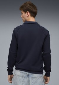 Maglione blu navy con collo e maniche lunghe, con polsini a coste. Sul retro presenta dettagli decorativi lungo le spalle. Tessuto morbido e strutturato.