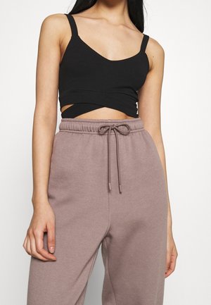 Vrouw draagt een zwart cropped tanktop met uitsnijdingsdetail en mauve hoge taille joggingbroek met trekkoord, staand tegen een witte achtergrond.
