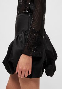 Minissaia preta de cetim com forma volumosa e manga comprida em malha texturizada adornada com lantejoulas pretas e prateadas em padrões florais.