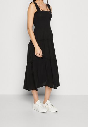 Schwarzes, gestuftes Midi-Kleid mit Rüschen-Trägern und gerafftem Oberteil, kombiniert mit weißen Sneakers. Der Stoff wirkt leicht und hat eine glatte Textur.