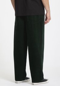 Groene corduroy wijduitlopende broeken met verticale ribbels, voorzien van achterzakken, gedragen met zwarte canvas sneakers. De textuur lijkt zacht en luxueus.
