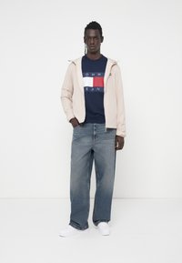 Šviesiai smėlinė užtrauktuku užsegama striukė, tamsiai mėlynas džemperis su raudonu ir baltu logotipu, plačios mėlynos džinsinės kelnės ir baltos sportinės bateliai. Paprastas ir kasdieniškas dizainas.