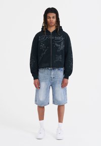 Felpa nera con zip e design grafici, abbinata a pantaloni di jeans denim chiari e sneakers bianche, indossata da una persona in piedi.