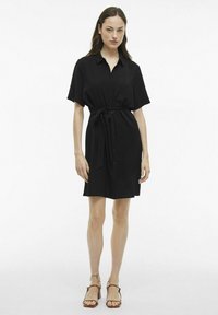 Juodas trumpas suknelė su marškinių coloru, mygtukų priekyje ir juostos raišteliais juosmenyje. Lygi tekstūra ir iki kelių ilgio dizainas, derinamas su rudos spalvos šlepetėmis.