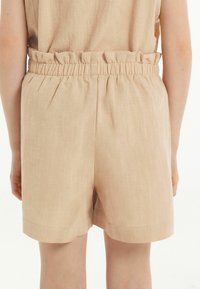 Pantaloni corti beige realizzati in tessuto leggero con vita elastica e orlo arricciato. L'orlo è dritto e finisce a metà coscia.